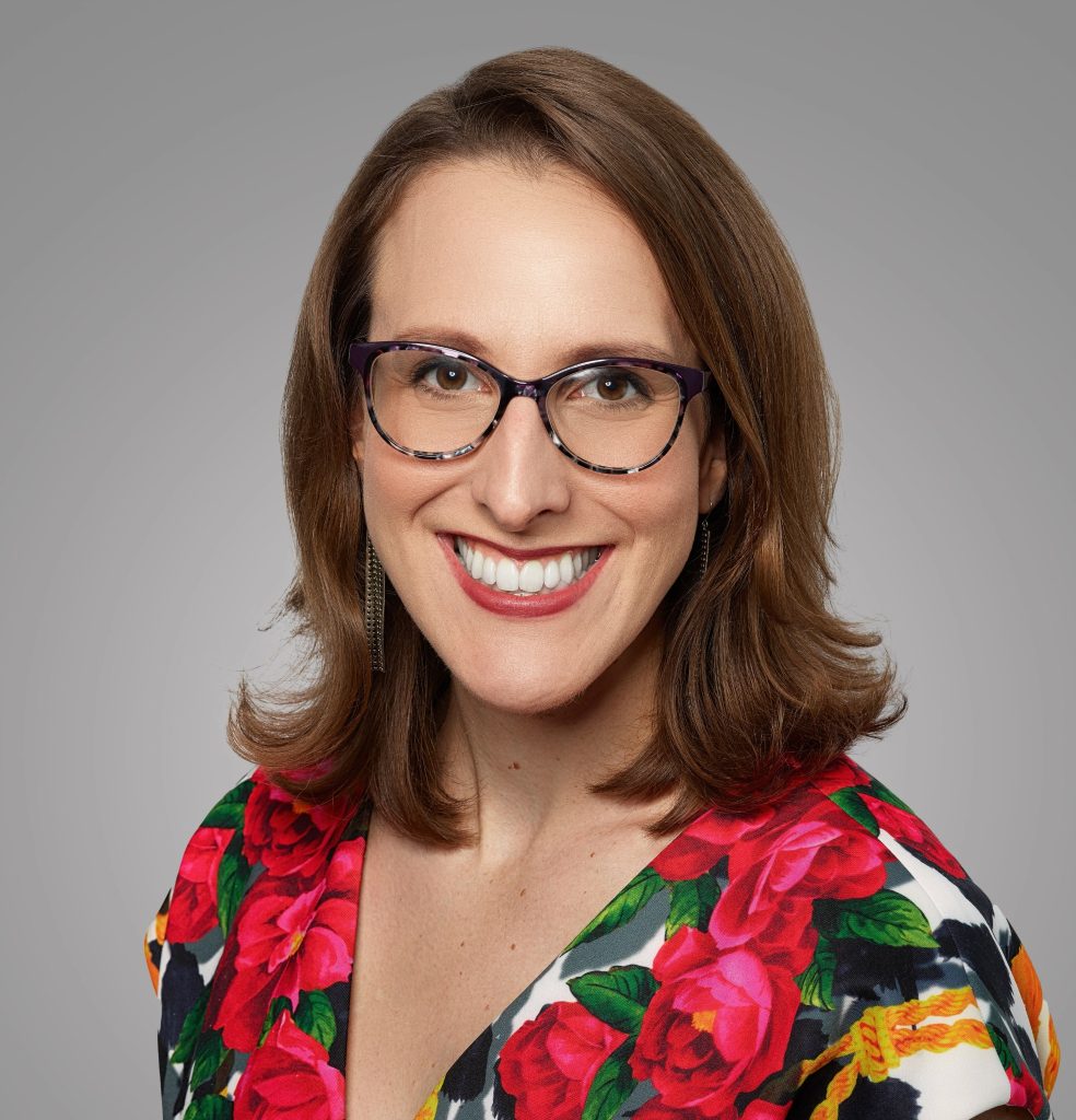 Headshot of Ashley Rose Young smiling at the camera in glasses and a colorful floral top against a gray background.