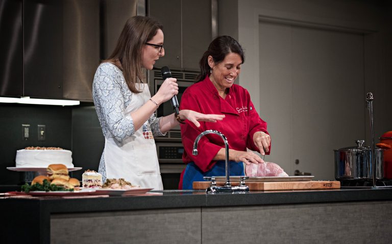 Ashley Rose Young speaks into a microphone beside a guest in a red shirt during a cooking demonstration. They stand behind a counter with food and cooking equipment.